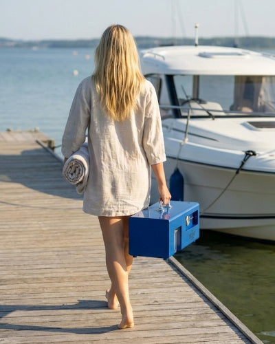 Woman walking on a dock with a blue cooler and towel, near a boat.